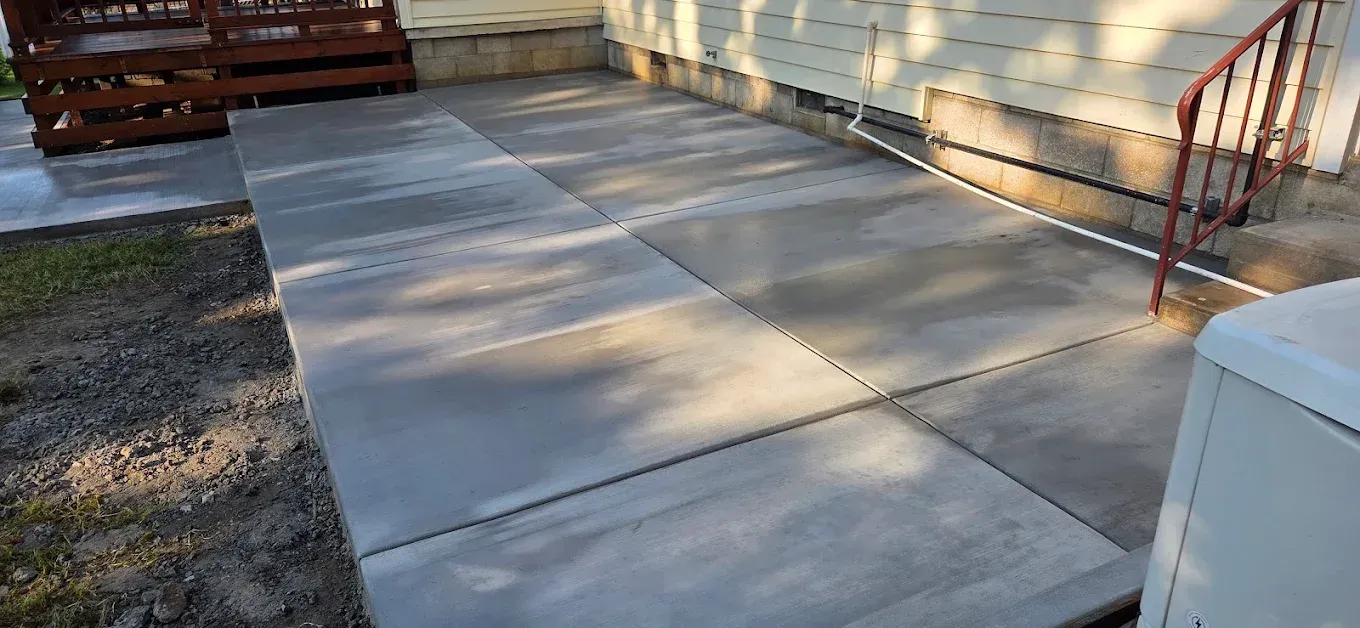 Modern home exterior with gray siding, large windows, and a covered patio with stone pillar and paved area.