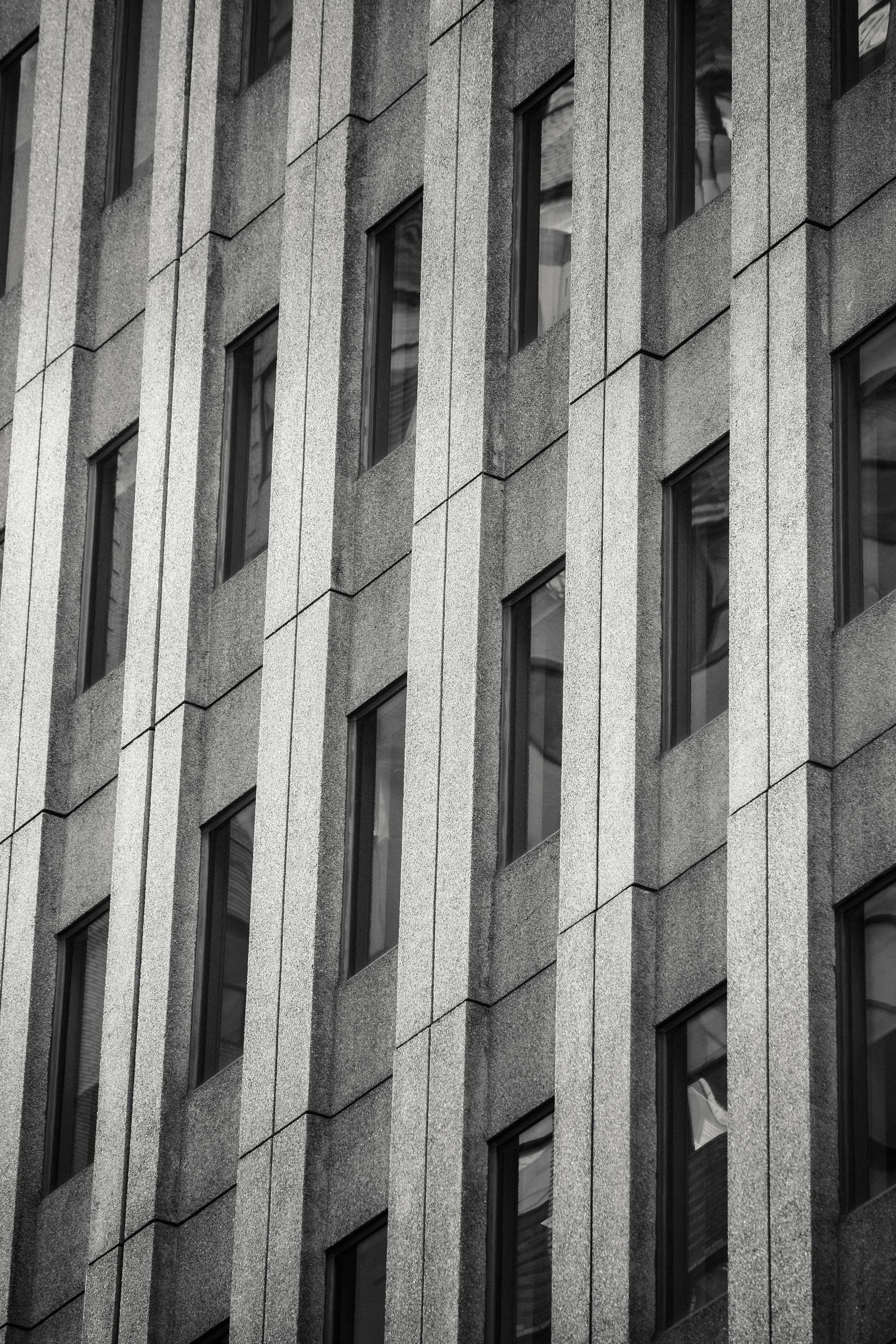 Black and white photograph of a tall building exterior, patterned with vertical columns of windows and textured concrete.