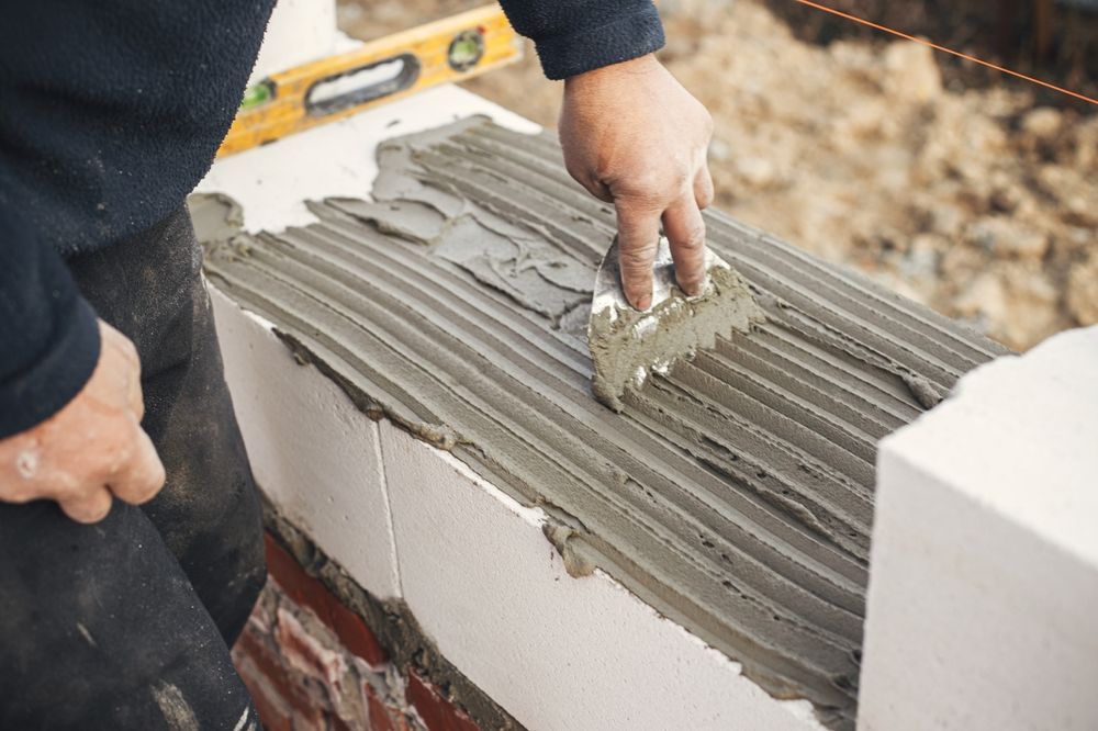 Person applying mortar with a notched trowel to a concrete block in an outdoor construction setting.