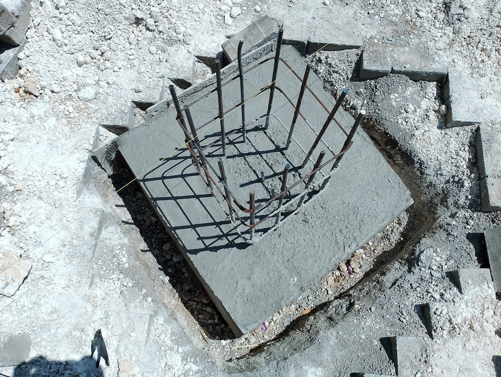Square concrete foundation with exposed rebar cage, set in a carved out area, surrounded by concrete blocks.