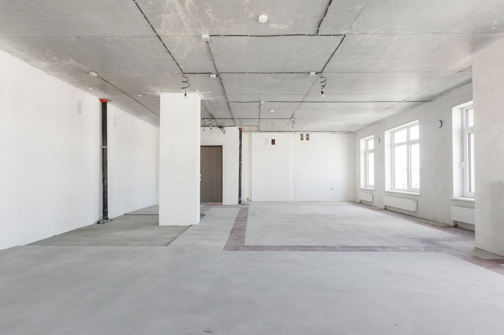 Empty, unfinished interior space with white walls, concrete floor, exposed wiring, and windows.