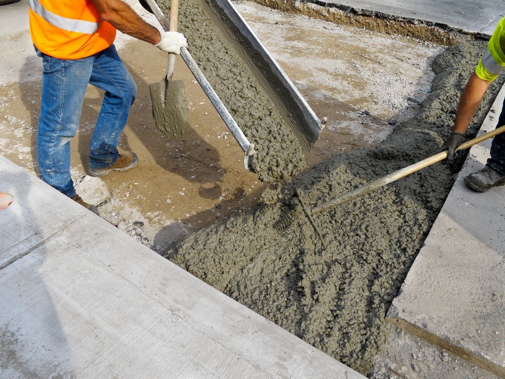 Workers pouring concrete into a form on a sidewalk. One wears orange safety vest.