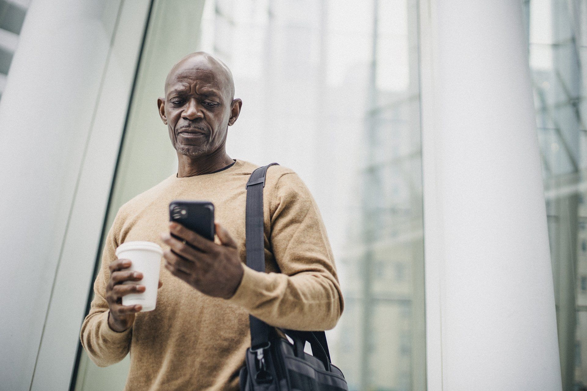 Man holding phone and coffee, looking at screen. Outdoors, modern building background.