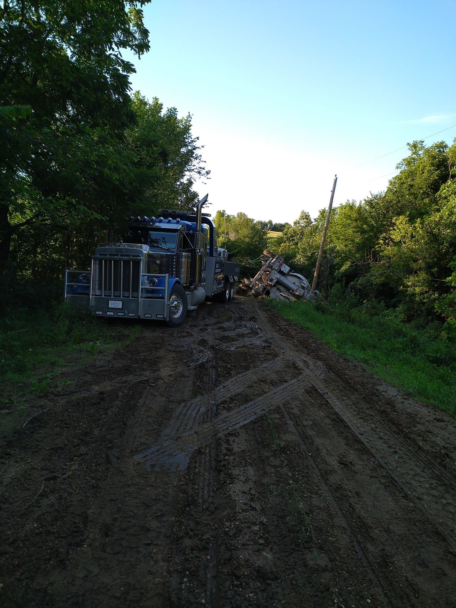 a heavy-duty wrecker assists an overturned semi truck on a dirt road surrounded by trees