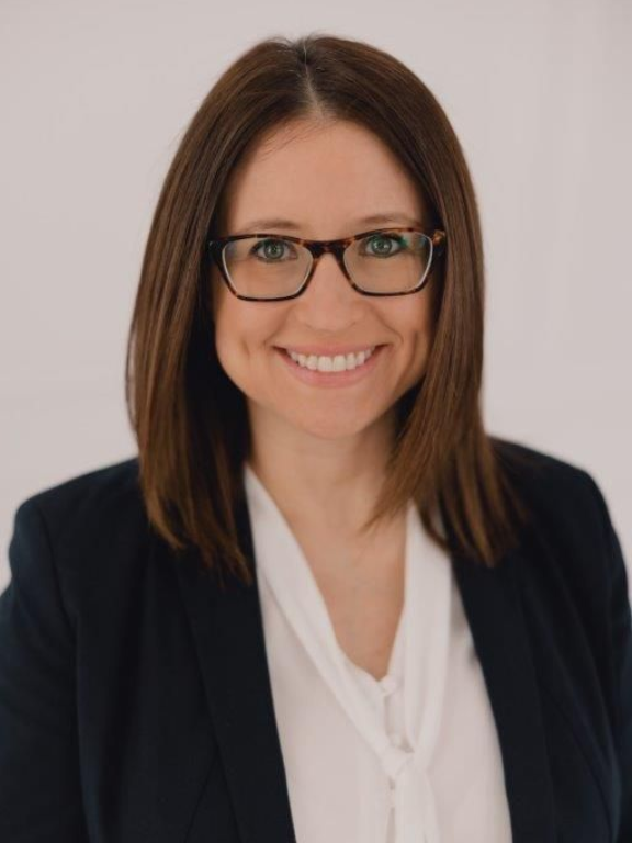 Woman with brown hair and glasses smiling, wearing a white shirt and navy blazer.