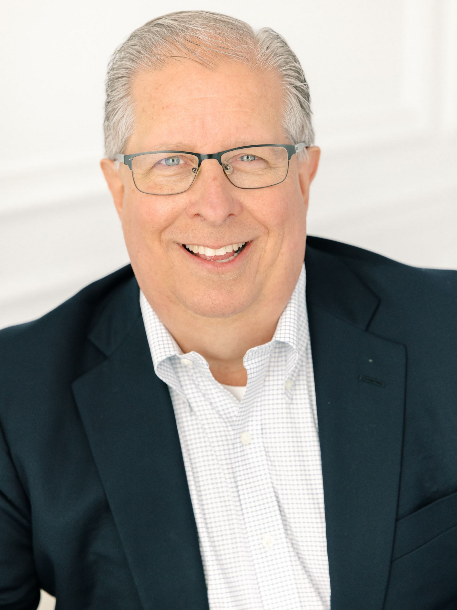 Man with glasses smiles, wearing a navy blazer over a light blue collared shirt.