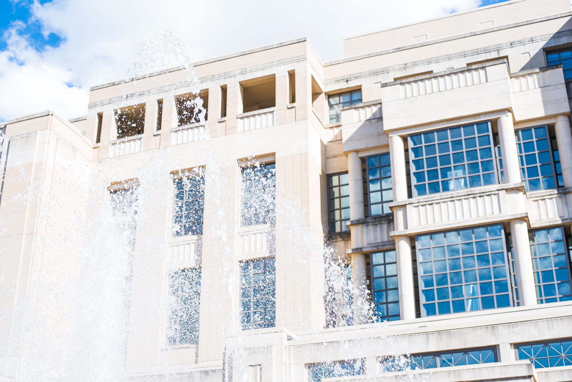 Beige building with blue-tinted windows, partly obscured by a fountain's water spray, against a cloudy sky.