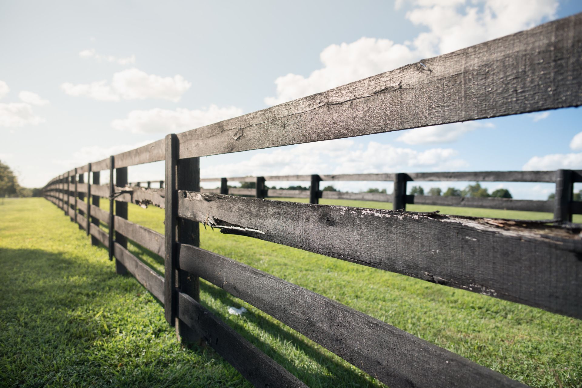 Black wooden fence in a green field under a partly cloudy blue sky.