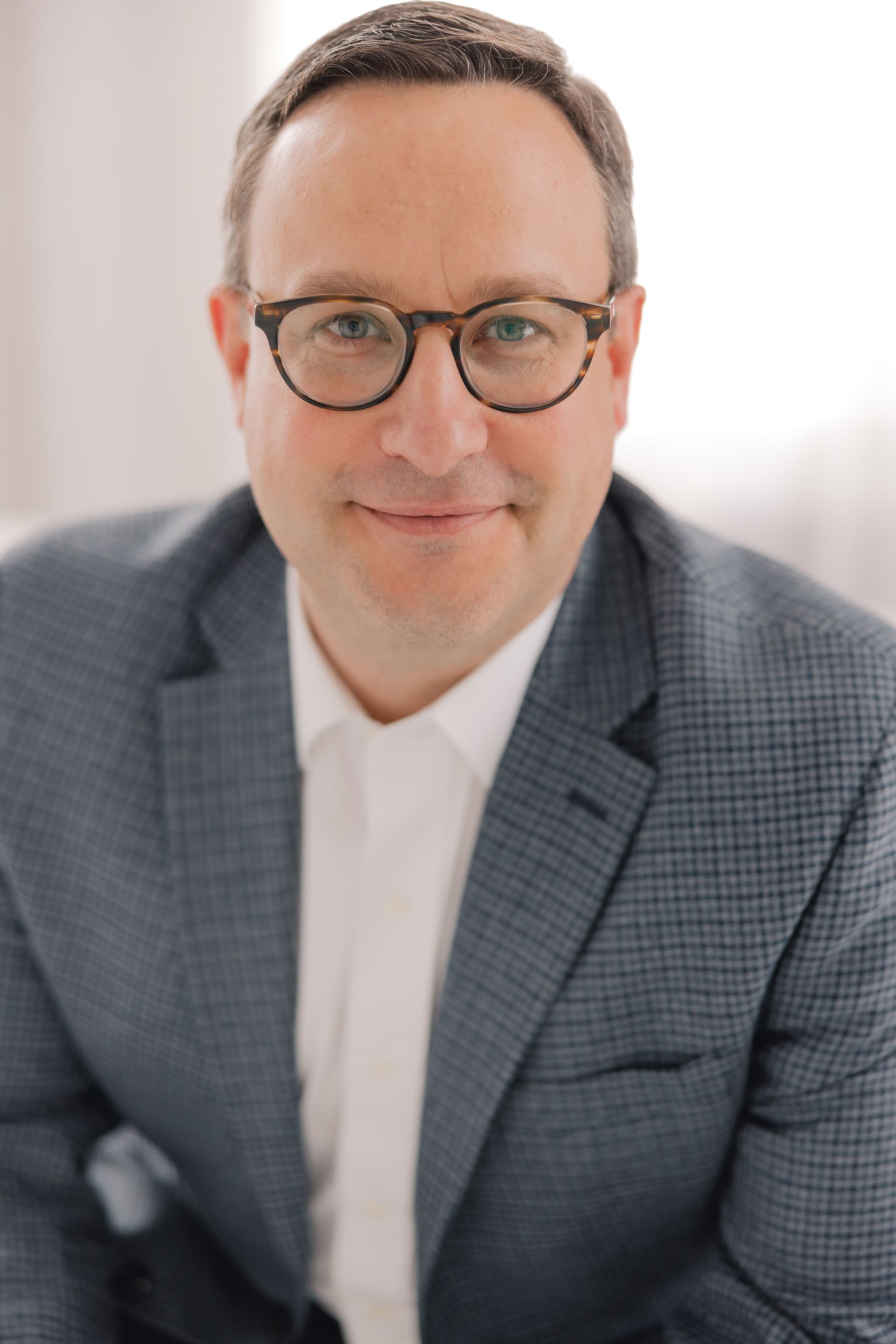 Man wearing glasses, blazer, and white shirt smiles at the camera, a blurred background.