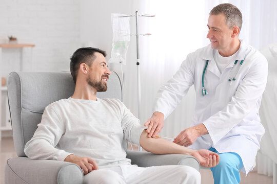 Man receiving IV fluids, sitting in an armchair, doctor attending. White room, medical equipment present.