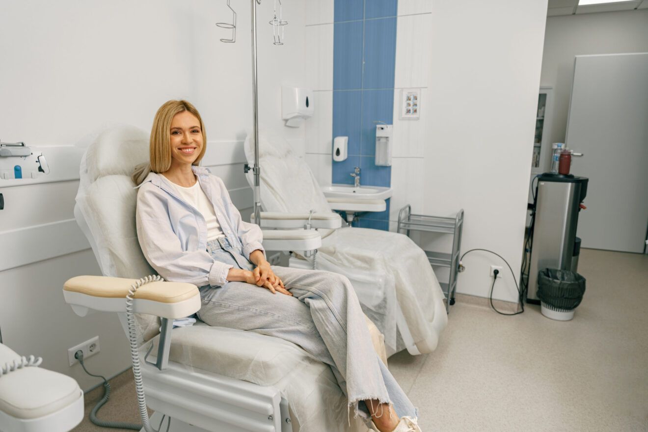 Woman sitting in medical chair, smiling, with IV drip in a medical room.
