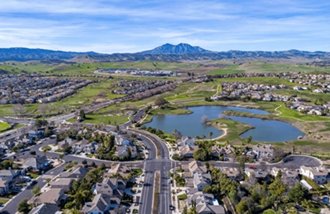 Aerial view of a suburban neighborhood with a lake, road, and mountain backdrop under a blue sky.