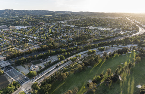Aerial view of suburban area with roads, trees, and a golf course, bathed in sunlight.