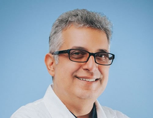 Man with glasses smiles, wearing a white coat against a blue background.