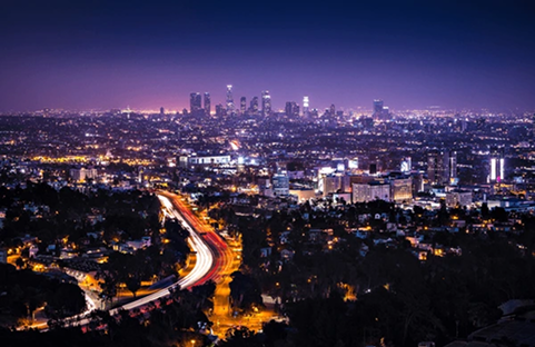 Night view of Los Angeles skyline with city lights, freeway streaks, and purple-blue sky.