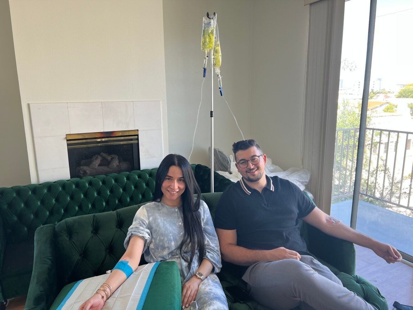 Two people receive IV drips on a green couch. A drip stand is between them, sunlight floods the room.