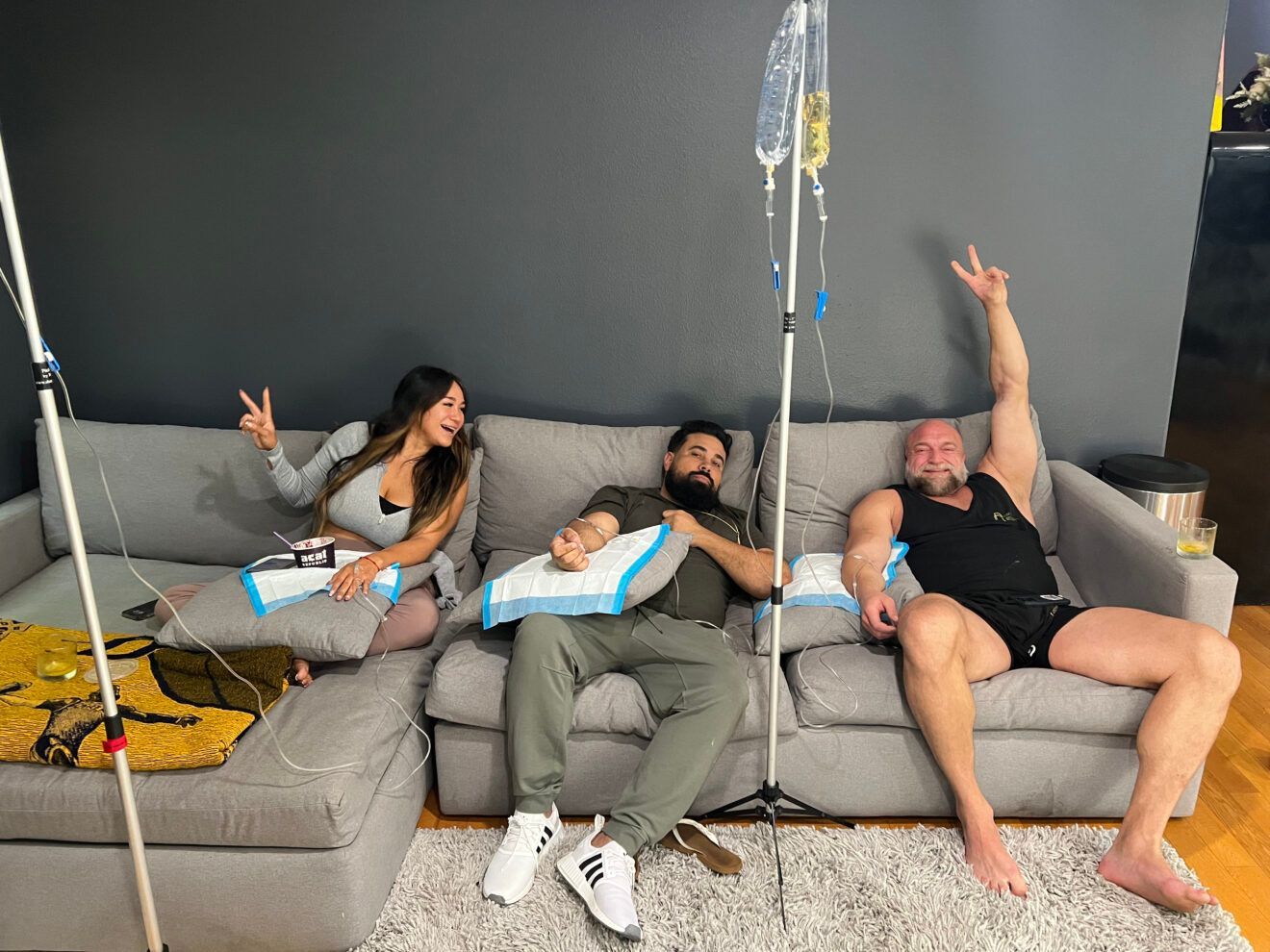 Three people reclining on a gray sofa, receiving IV drips. They appear relaxed, making peace signs. Dark room.