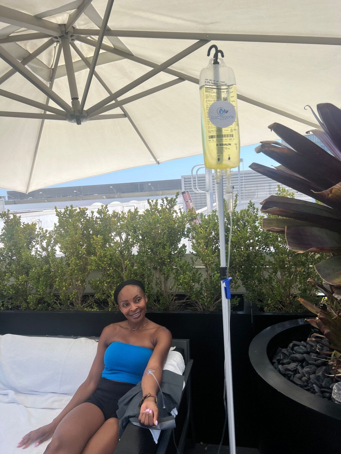 Woman receiving IV therapy outdoors, smiling, under a white umbrella near plants.