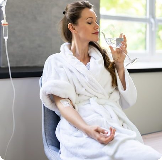 Woman in a robe receiving IV fluids, drinking water, seated near window.