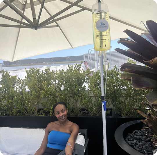 Woman receiving IV fluids outdoors, smiling. Blue top, under white umbrella, with greenery.