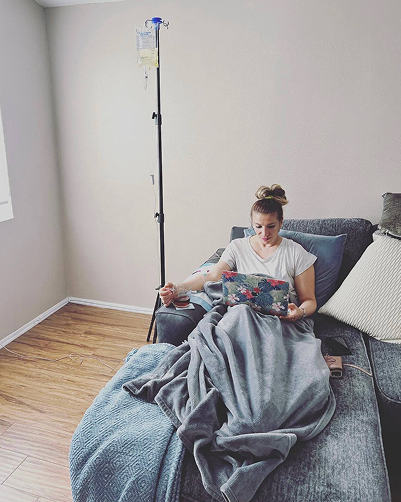 Woman receiving IV fluids on a couch, reading. Dark-haired, casual clothes, blanket, light room, and IV stand.