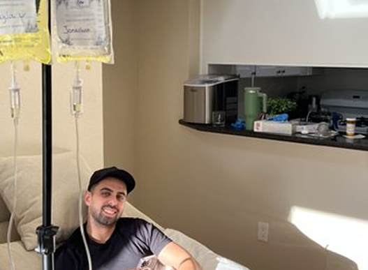 Man receiving IV treatment, smiling while relaxing on a couch. IV bags and stand visible.
