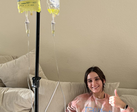 Woman seated with IV drip, giving thumbs up, smiling, on a beige couch.