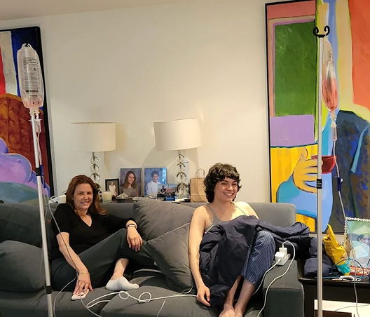 Two women on a sofa receiving IV drips in a brightly colored room. Smiling.