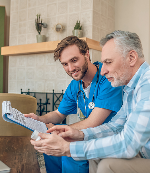 Male nurse in blue scrubs reviewing medication with an older man in a living room.