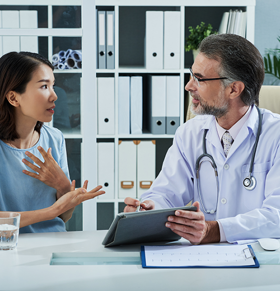 Woman talking to a doctor in an office; she gestures with hands. The doctor holds a tablet.