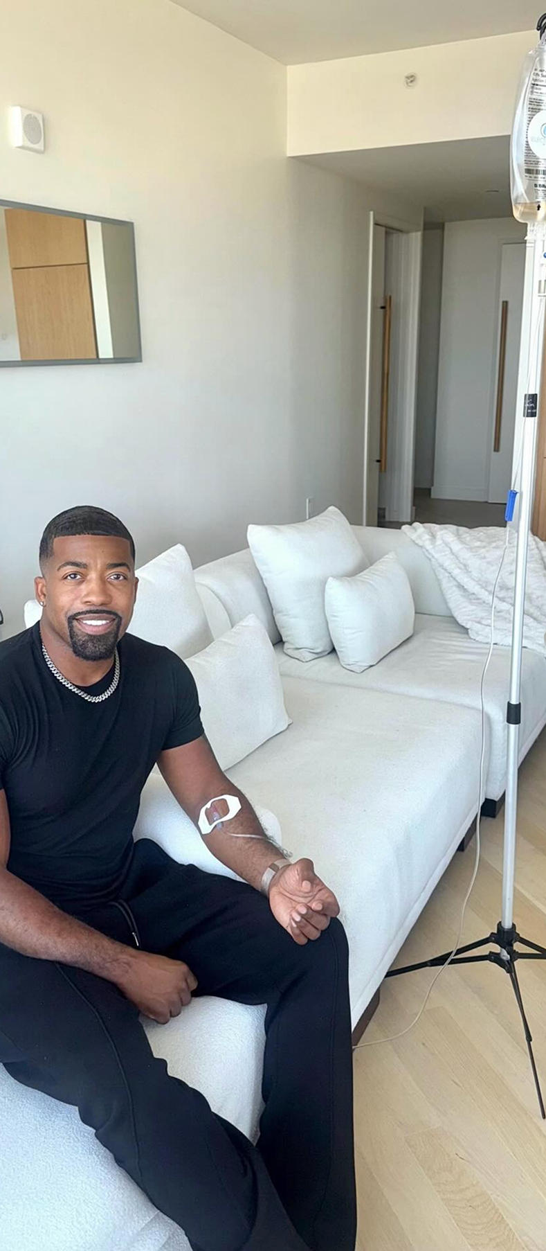 Man sitting on couch receiving IV drip. He is wearing black clothes. White couch in a modern room.