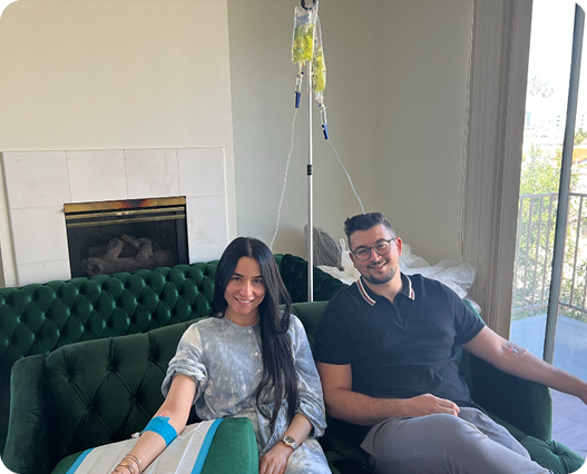 A woman and man seated on a green couch, receiving IV drips in a living room.