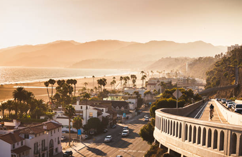 Coastal town view at sunset with ocean, beach, palm trees, road, bridge, and mountains.