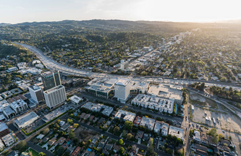 Aerial view of a cityscape with buildings, a highway, and lush green trees under a warm sunset glow.