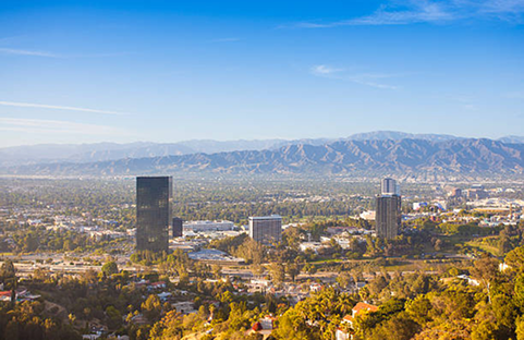 Cityscape with tall buildings, trees, and mountains under a blue sky.