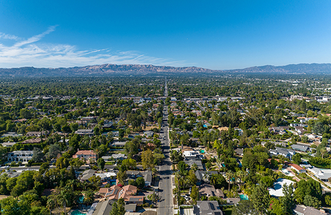 Aerial view of a suburban neighborhood with houses, trees, and a long road extending to mountains under a blue sky.