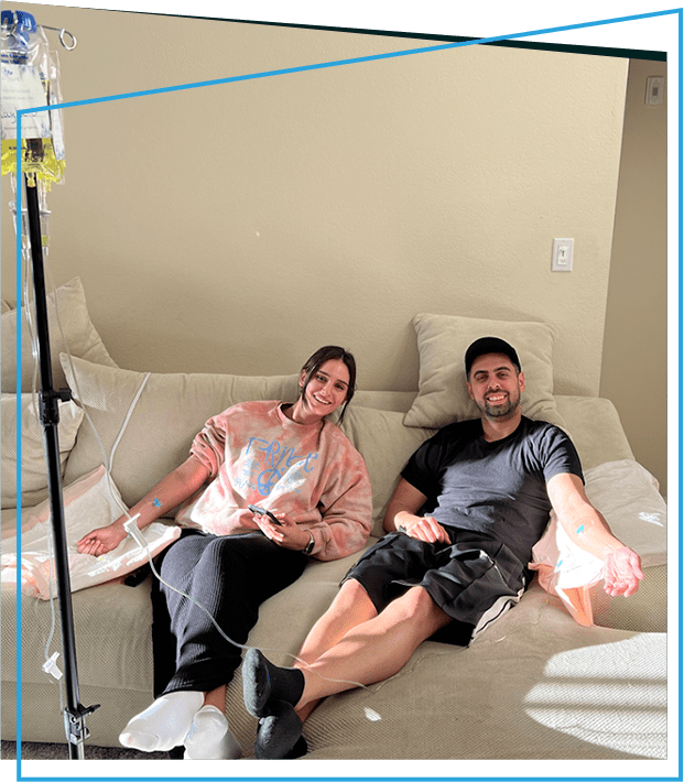 Two people on a couch receiving IV fluids; light-skinned, smiling, in casual clothes.