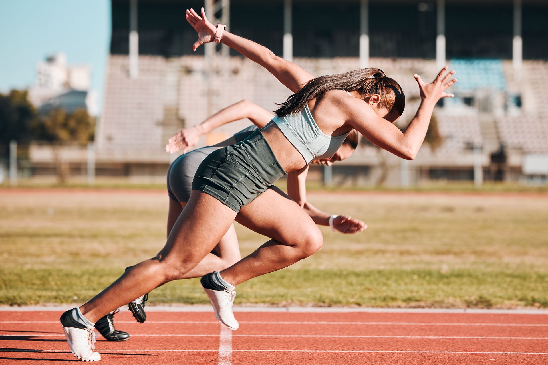 Two sprinters in athletic wear take off from starting blocks on a red running track at an outdoor stadium.