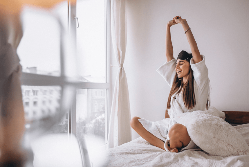 Woman in robe stretching on a bed near a window, waking up.