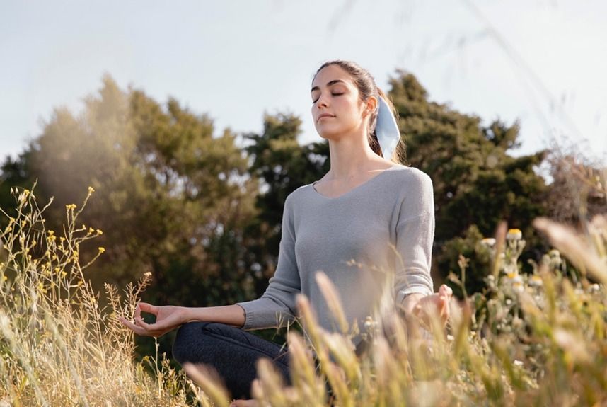 Woman meditating outdoors in a field, eyes closed, wearing gray top and blue hair tie, hands in lotus position.