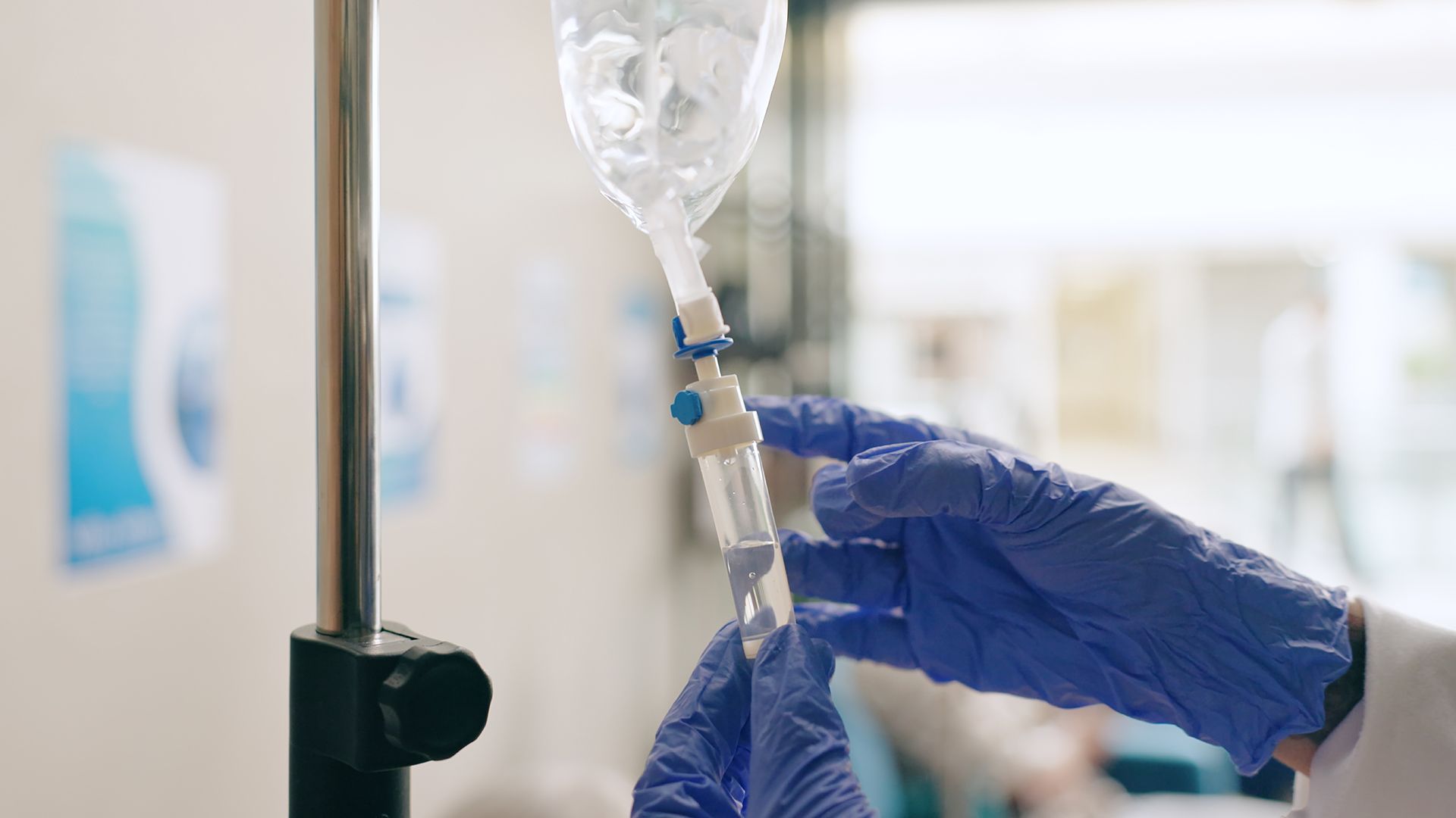 A medical professional in blue gloves adjusts the drip rate on an IV bag in a clinical setting.