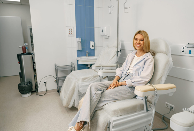 Woman in medical chair smiling in a clinical setting with IV stand and medical equipment.