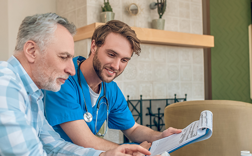 A healthcare provider shows paperwork to an older adult patient, both smiling in a living room.