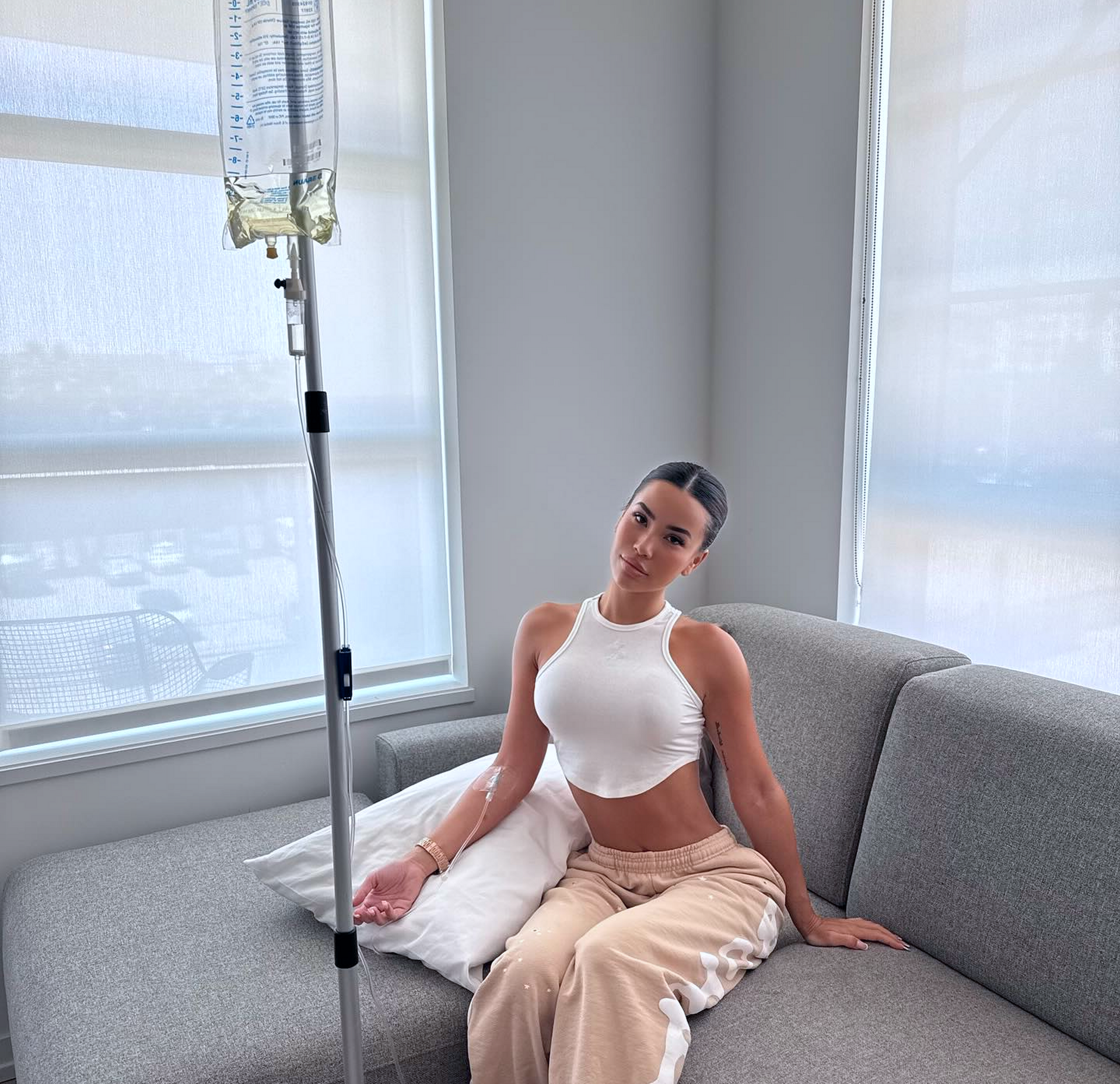 Woman on a couch receiving IV fluids; she is smiling, wearing a white top and tan pants. Bright room.