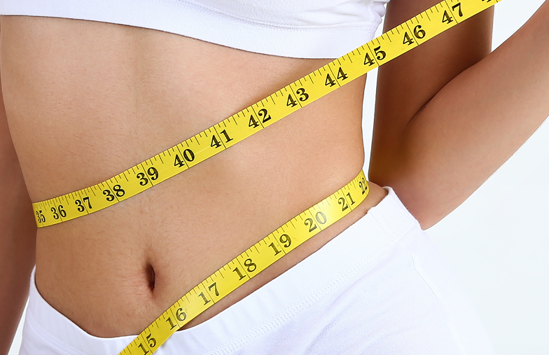 Person's waist measured with yellow tape. Wearing white top and pants.