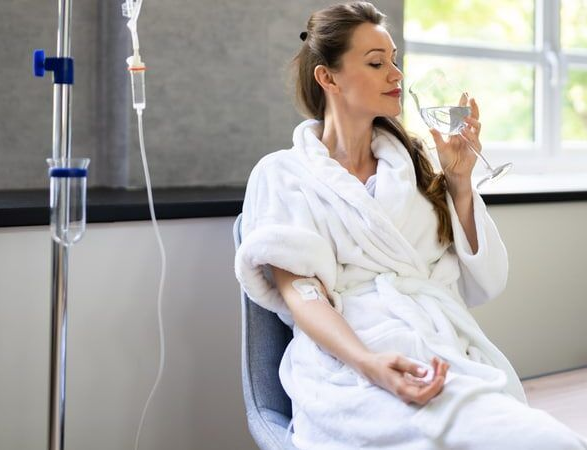 Woman in a robe with IV drip in arm drinks water, seated indoors near window.