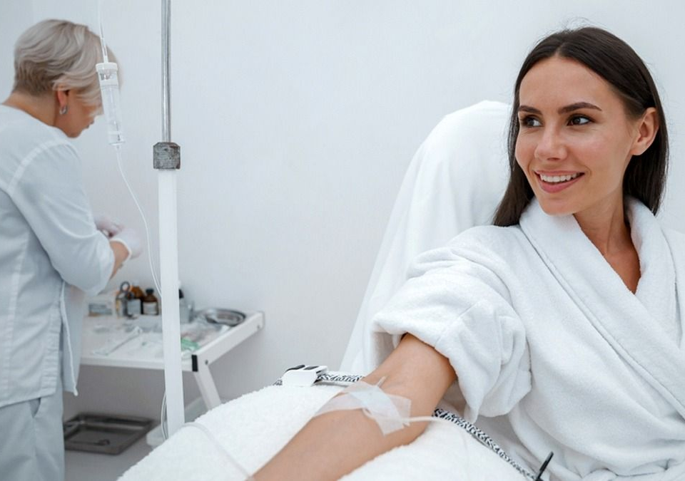 Woman in white robe receiving IV treatment; nurse preparing medication in white room.