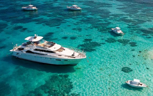 An aerial view of a large white yacht anchored in clear, turquoise tropical waters with several smaller boats nearby.