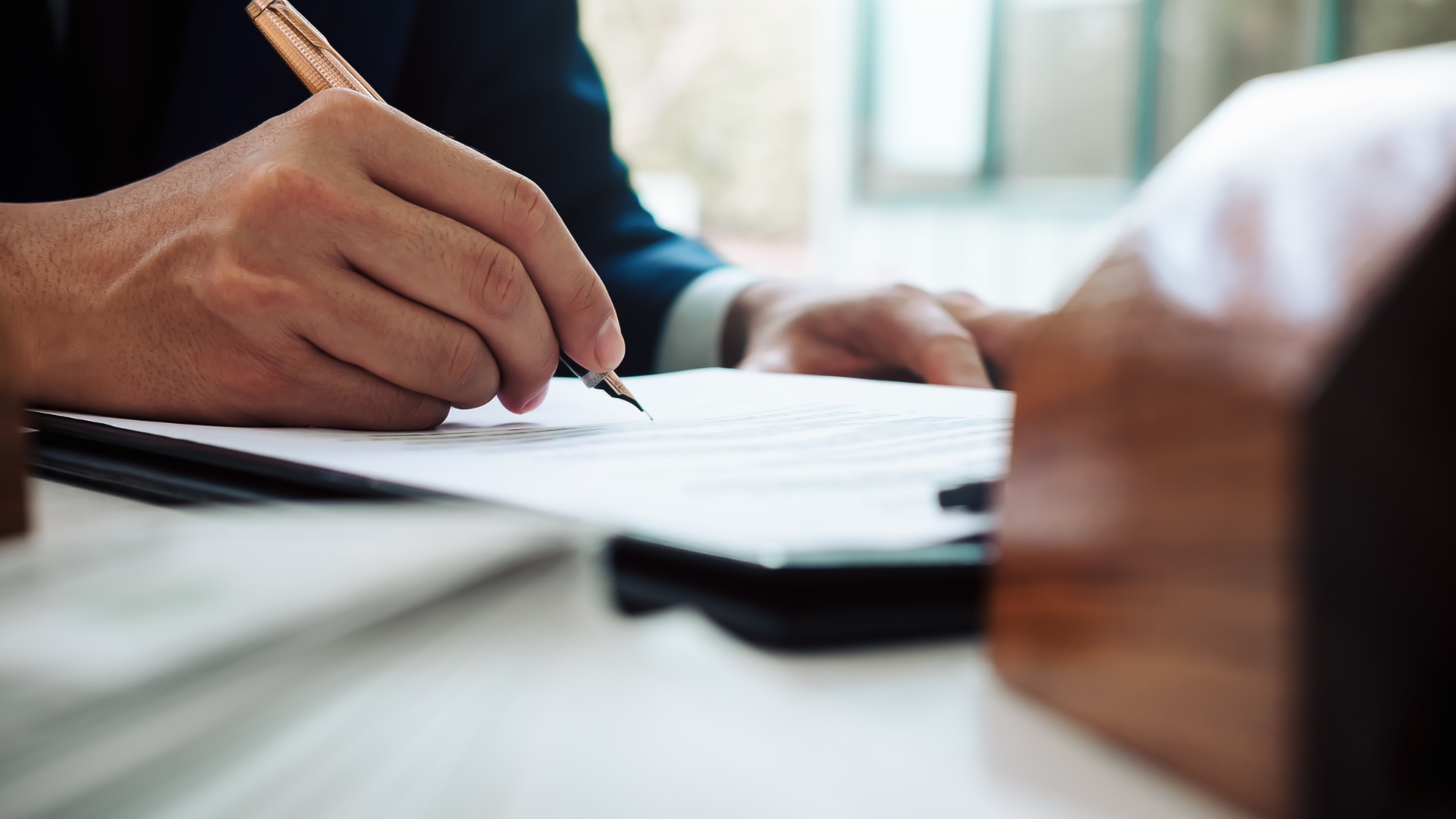 Person in a suit signing a document with a pen; a small wooden house visible on the right.