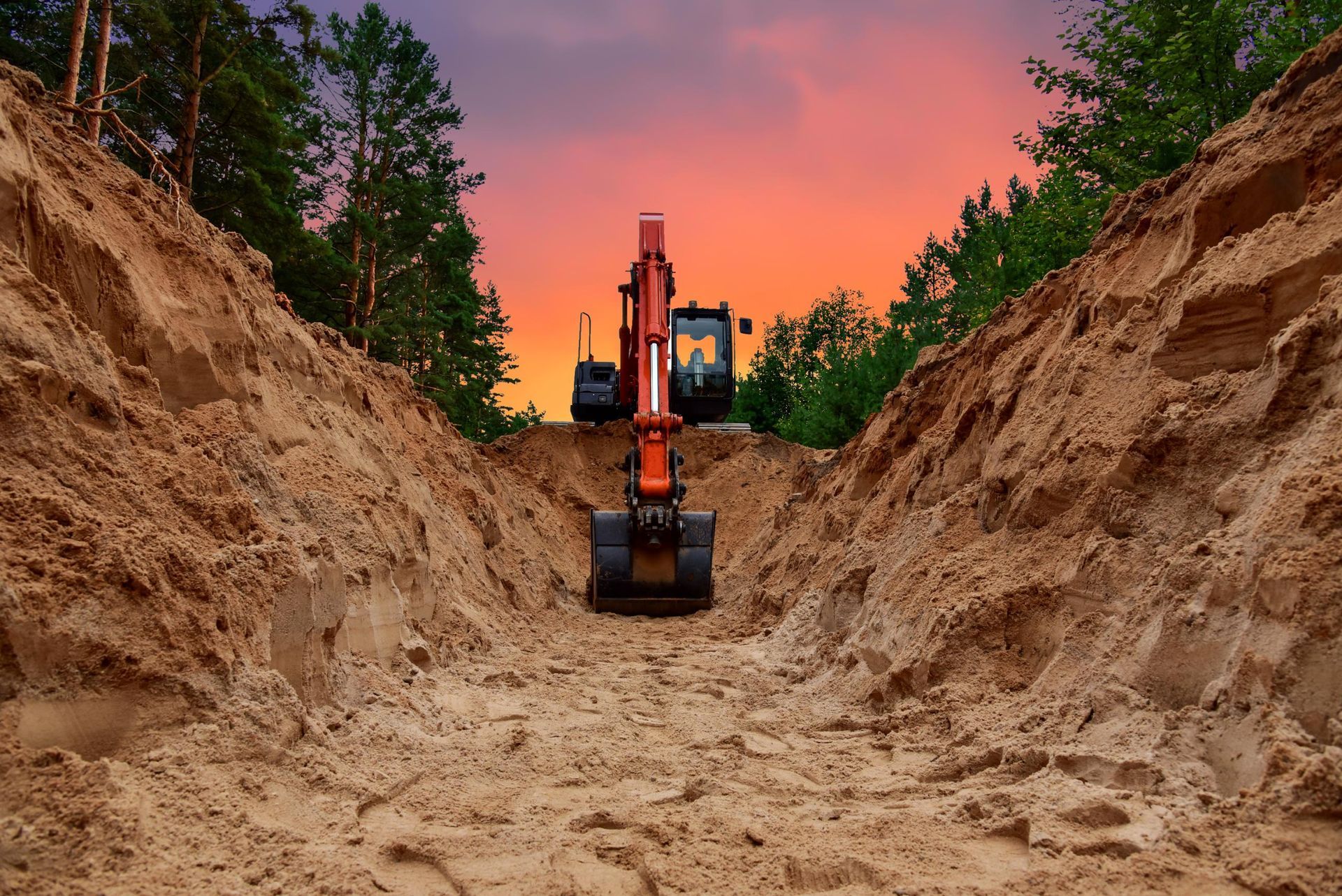 Excavator digging a dirt trench at sunset, framed by steep earth walls and trees under a pink sky